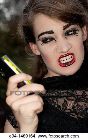Picture - A 19 year old brunette woman with dramaitc make up showing her teeth to a mobile phone.. Fotosearch