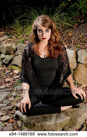 A 19 year old brunette woman with dramaitc make up looking directly at the camera sitting cross legged on rocks in a garden. View Large Photo Image Stock Photo - A 19 year old brunette woman with dramaitc make up looking directly at the camera sitting cross legged on rocks in a garden.. Fotosearch