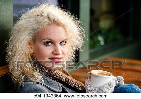 Picture - A 22 year old blond woman sitting at an outdoor cafe with a cup of coffee in the winter.. Fotosearch