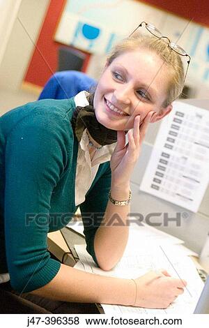 Stock Photo - A 23 year old girl sitting at a desk working looking off camera smiling. Fotosearch