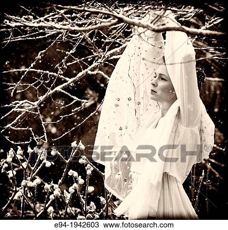 Stock Image - A 24 year old woman wearing a white dress standing in a garden during a snow storm.. Fotosearch