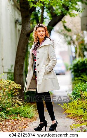 A 27 year old brunette woman wearing a white winter coat, black leggings and high heal shoes standing on a path way between 2 buildings. View Large Photo Image Picture - A 27 year old brunette woman wearing a white winter coat, black leggings and high heal shoes standing on a path way between 2 buildings.. Fotosearch