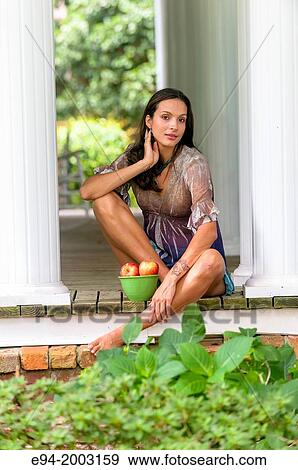 Stock Photo - A 27 year old brunette woman wearing a short summer dress sitting on a porch with a bowl of apples.. Fotosearch