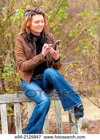 Stock Photo - A 27 year old brunette woman wearing a brown leather jacket sitting on the back of a wooden bench looking at her cell phone.. Fotosearch