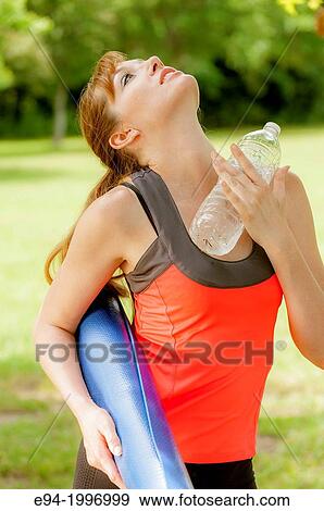 Stock Photo - A 31 year old red headed woman in work out clothing holding a yoga mat and a bottle of watter in a park setting.. Fotosearch