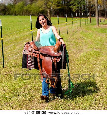 Stock Photography - A 36 year old brunette woman carrying a saddle in a pasture.. Fotosearch