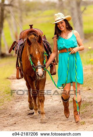 A 36 year old brunette woman weaing a dress and cowboy boots and straw hat leading a horse on a farm. View Large Photo Image Stock Photograph - A 36 year old brunette woman weaing a dress and cowboy boots and straw hat leading a horse on a farm.. Fotosearch