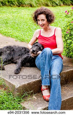 Stock Photograph - A 51 year old brunette woman wearing jeans sitting on steps in front of her house playing with her dog.. Fotosearch