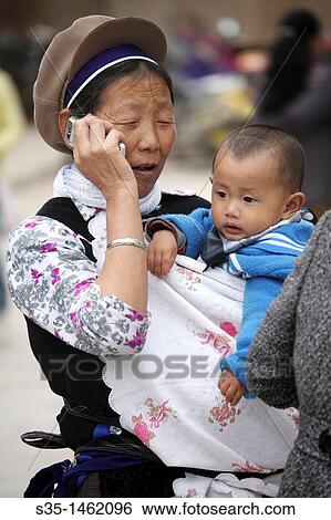 Stock Photograph - A Bai nationality lady carrying a baby and talk on a cell phone Kunming China.. Fotosearch