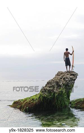 Stock Photography - A Balinese surfer perched on top of a rock looking out over the ocean. Fotosearch