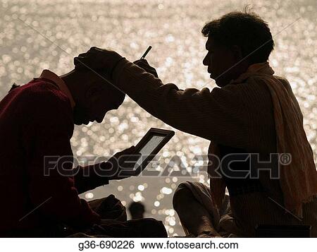 Stock Photograph - A barber shaves a man's head with a straight razor in the early morning sun on the banks of the Ganga river in Varanasi, India. Fotosearch