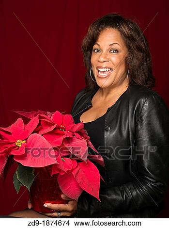 Picture - A beautiful middle-aged black woman beams with delight as she holds a poinsettia. Fotosearch