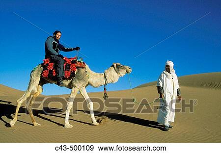 Stock Photo - A Berber at Tinfou - photogenically dressed up as an Arab nomad - takes a tourist on a camel ride into the dunes. Dr? a valley, southern Morocco.. Fotosearch