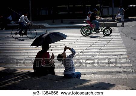 A boy drinks yogurt next to his mother holding an umbrella to shade them from the sun as they watch cyclists ride along Paseo de la Reforma Avenue in Mexico City, November 27, 2009 View Large Photo Image Stock Photograph - A boy drinks yogurt next to his mother holding an umbrella to shade them from the sun as they watch cyclists ride along Paseo de la Reforma Avenue in Mexico City, November 27, 2009. Fotosearch