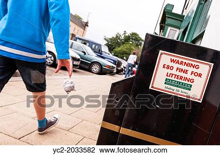 Stock Image - A boy throws litter on the ground as he walks past a bin warning of an? 80 fine for littering in the Uk.. Fotosearch