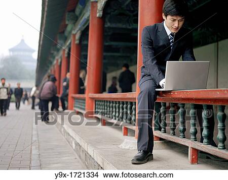 A Chinese businessman on his laptop while sitting on traditional hand railing. View Large Photo Image Picture - A Chinese businessman on his laptop while sitting on traditional hand railing.. Fotosearch
