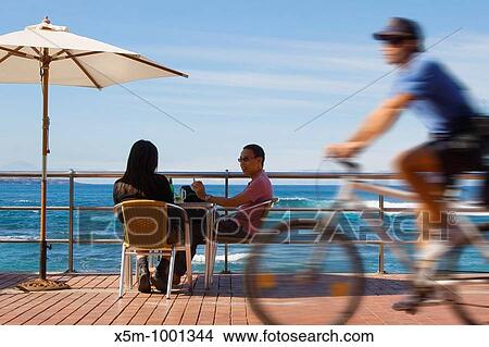 A couple sit at a beachfront table on Las Canteras Beachfront as a policeman cycles past View Large Photo Image Picture - A couple sit at a beachfront table on Las Canteras Beachfront as a policeman cycles past. Fotosearch