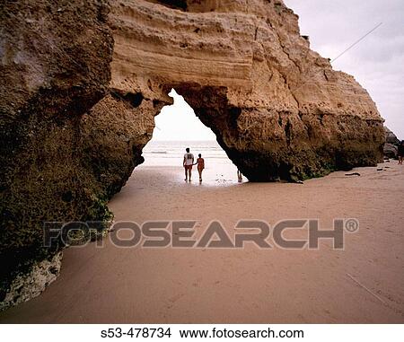Picture - A couple strolls under arch on beach in Algarve. Fotosearch