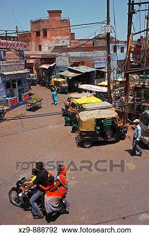 Stock Image - A four-members Indian family on a motorbike, people walking around and some rickshaws parked on a little square in Agra. Fotosearch