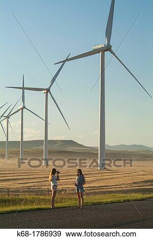 Stock Photo - A girl, 16, takes a photo of her sister, 20, near wind turbines near Pincher Creek in southern Alberta, Canada. Fotosearch
