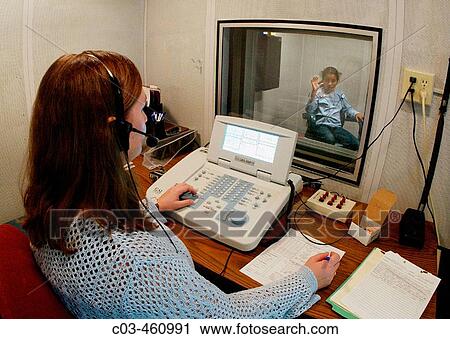 Stock Image - A hearing impaired adolescent Hispanic girl in a soundproof testing room signals to an audiologist that her hearing aid is functioning correctly at a speech and hearing clinic in Orange, California. Fotosearch