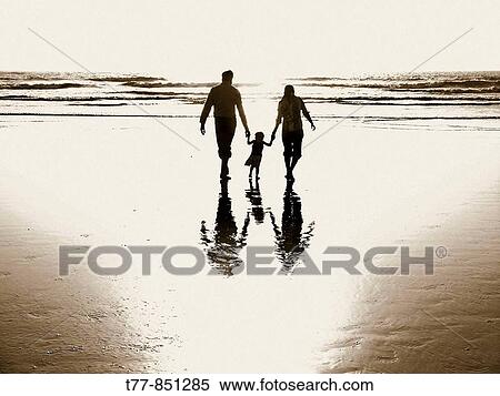 Stock Photography - A husband and wife, holding the hands of their baby girl, helping her walk with them through the ocean surf. Fotosearch