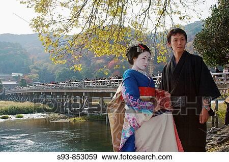 Stock Photo - A Japanese man wearing a traditional kimono and a woman dressed up as a geisha are standing infront of the famous bridge Togetsu-kyo, the dominant landmark in Arashiyama with autumn leaf colors in the back ground. Fotosearch