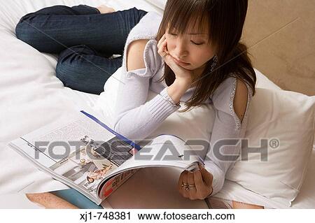 Stock Image - A Japanese woman resting on her hotel room bed reading a magazine. Fotosearch