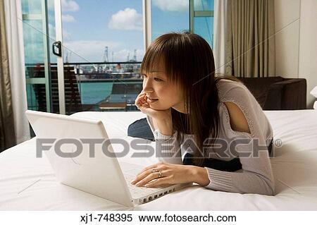 Stock Photography - A Japanese woman resting on her hotel room bed using a laptop computer.. Fotosearch