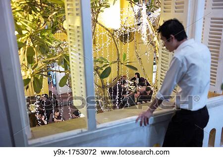 Stock Image - A man looking down at the interior of Nha Hang Ngon restaurant with lights on trees. Fotosearch
