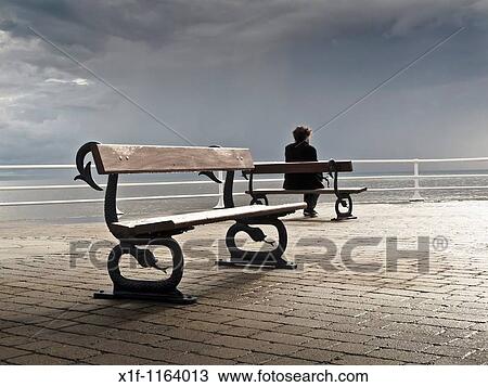Stock Image - A man sitting alone on a bench on the promenade, grey overcast July summer afternoon, Aberystwyth Wales UK. Fotosearch