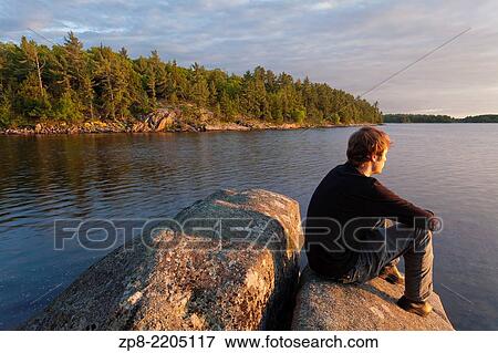 Stock Photo - A man sitting on a rock as the sun sets over Georgian Bay. Massasauga Provincial Park, Ontario, Canada.. Fotosearch