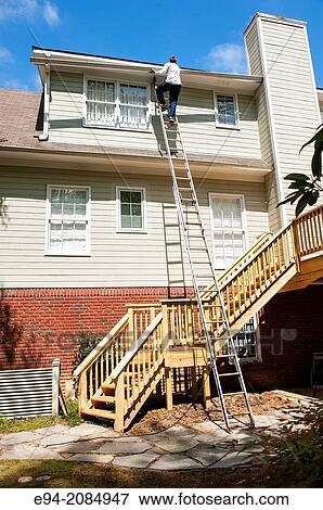 A man standing on a ladder on the side of a house lifting a drain gutter. View Large Photo Image Stock Photo - A man standing on a ladder on the side of a house lifting a drain gutter.. Fotosearch