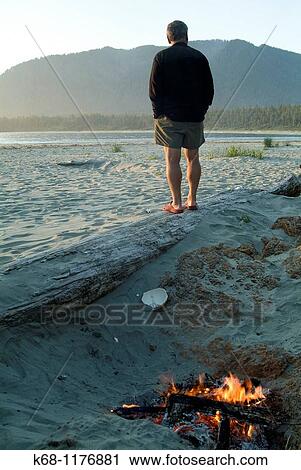 Stock Image - a man stands on a beach by a campfire on Flores Island, Clayoquot Sound, British Columbia, Canada. Fotosearch