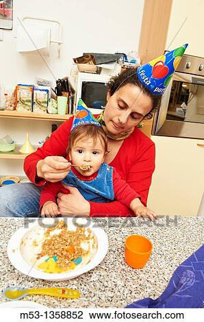 A mother feed her 10 months baby girl in the family kitchen Both wear party hats View Large Photo Image Stock Image - A mother feed her 10 months baby girl in the family kitchen Both wear party hats. Fotosearch