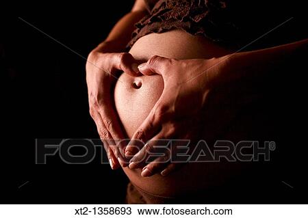 Stock Image - A pregnant woman makes a heart shape over her pregnant stomache on a black background. Fotosearch