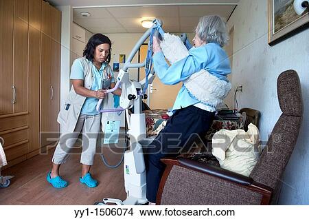 Picture - A professional nurse of a Rotterdam nursing home helping an elderly inhabitant from her chair, using a lifter.. Fotosearch
