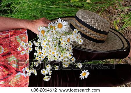 Stock Image - A senior woman carrying a wide brimmed hat and a bouquet of daisies in the garden. Fotosearch