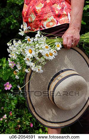 Stock Image - A senior woman carrying a wide brimmed hat and a bouquet of daisies in the garden. Fotosearch