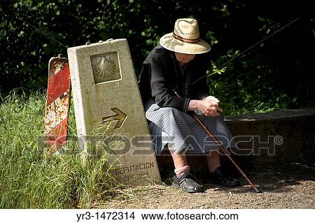 Picture - A senior woman rests next to a boundary stone in the French Way that leads to Santiago, in Casanova village, Galicia region, Spain.. Fotosearch