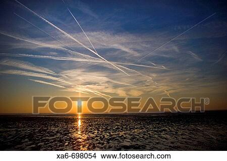 A setting sun creates an orange layer in the deep blue sky and blazes a gold trail across the wet sand of the beach. White vapour trails criss-cross the sky. View Large Photo Image Picture - A setting sun creates an orange layer in the deep blue sky and blazes a gold trail across the wet sand of the beach. White vapour trails criss-cross the sky.. Fotosearch