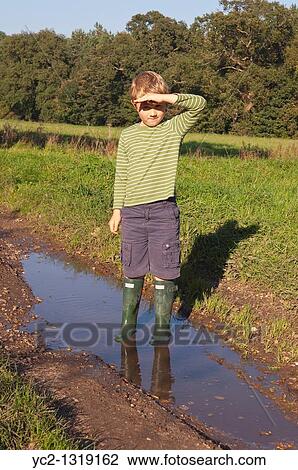 Stock Image - A seven year old boy stands in a puddle with Hunter wellies on in the Uk. Fotosearch