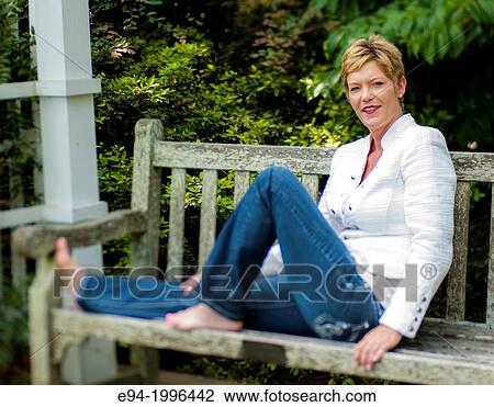 A smiling 44 year old blond woman wearing blue jeans and a white jacket sitting on a bench in a garden setting. View Large Photo Image Stock Image - A smiling 44 year old blond woman wearing blue jeans and a white jacket sitting on a bench in a garden setting.. Fotosearch