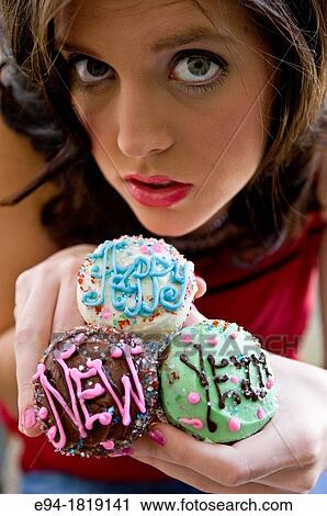 Stock Image - A tight portrait of a 19 year old brunette woman holding 3 cupcakes that say Happy New Year in the icing.. Fotosearch