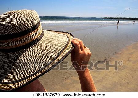 Stock Photo - a woman holding her hat while strolling on a beach. Fotosearch