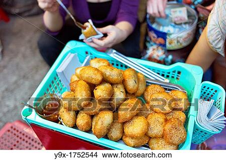 Picture - A woman selling the fried food banh ran, or deep fried rice ball, on the side of a street on makeshift displays and stools. Fotosearch