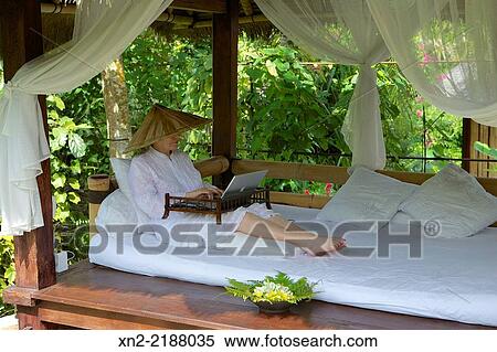 A woman uses a computer while lounging in an outdoor living room in the garden of a jungle home in Ubud, Bali, Indonesia View Large Photo Image Stock Photography - A woman uses a computer while lounging in an outdoor living room in the garden of a jungle home in Ubud, Bali, Indonesia. Fotosearch
