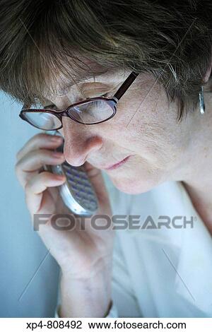 A woman wearing glasses and a white blouse listens on her mobile phone while looking downwards to a bright light source. View Large Photo Image Stock Image - A woman wearing glasses and a white blouse listens on her mobile phone while looking downwards to a bright light source.. Fotosearch