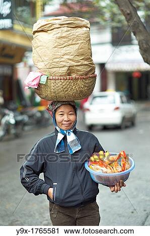 Stock Image - A woman with a large basket on her head selling bread and bananas on the streets of Vietnam. Fotosearch