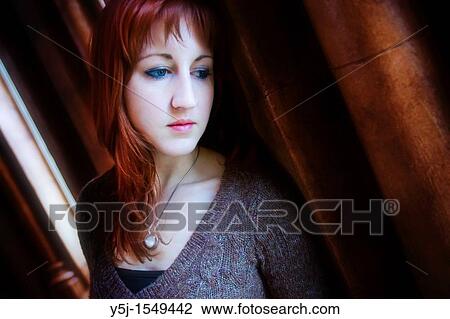 Stock Image - A youjng Caucasian female looking outside of window appearing melancholic and depressed. Fotosearch